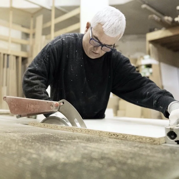 Un carpintero experimentado con gafas y pelo canoso operando una sierra de panel grande para cortar una pieza de madera con precisión en un taller de carpintería.
